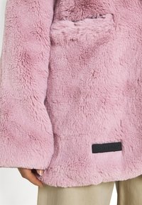 Roze nepbontjas met een zachte, textuurachtige oppervlakte. Beschikt over een borstzak en een zwart label met 'SIXTH JUNE' nabij de zoom.