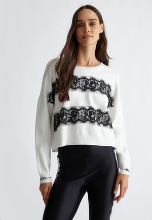 Weißer, kurzer Pullover mit schwarzen Spitzenakzenten und fransigen Kanten, kombiniert mit schwarzen Leggings, die mit einem weißen Streifen verziert sind.