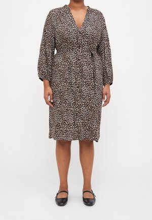 Kobieta w sukience w cętki sięgającej do kolan z długim rękawem i guzikami z przodu, nosząca czarne buty Mary Jane, stojąca na białym tle.