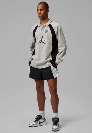 Homme debout les mains dans les poches, portant un polo gris et noir à manches longues, un short noir, des chaussettes blanches et des baskets Nike argentées sur un fond uni.