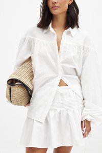 Chemise blanche à manches longues avec des manches froncées, associée à une mini jupe texturée blanche. Tient un sac à main en paille rond avec des accents noirs.