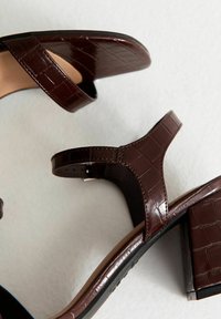 Sandalias de tacón cuadrado de piel de cocodrilo en tono marrón oscuro con correas en el tobillo sobre un fondo claro.