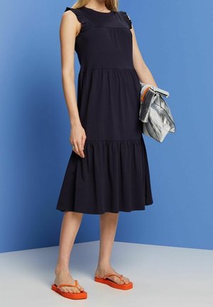 Frau in einem ärmellosen schwarzen Midikleid mit Rüschen an den Schultern, orangefarbenen Flip-Flops und einer großen silbermetallischen Clutch.