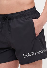 Czarne kąpielówki z elastycznym pasem, kieszenią z przodu i wiązaniem. Posiadają białe logo EA7 Emporio Armani z boku.