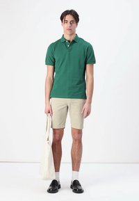 Žalias polo marškinėliai iš medvilnės, smėlio spalvos šortai, balti kojinės, juodi slip-on bateliai, laikantis smėlio spalvos rankinės. Paprastas dizainas, atsitiktinis kirpimas.