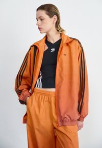 Oranžs zip-up jaku ar melnajām svītrām, apvienots ar melnu īso topu un atbilstošām oranžām biksēm. Gluda auduma ar brīvu piegriezumu.