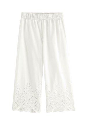 Smilende ung pige med langt blondt hår iført en stråhat, hvid blonderbluse og bukser samt mørke flettede sko, stående med hænderne på hofterne.