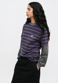 Prazna majica s prečkama v vijolični barvi z žepom, kombinirana s sivim dolgim rokavom spodaj. Obleka je dopolnjena s črnimi hlačami.