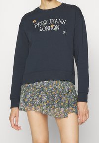 Felpa blu navy con "PEPE JEANS LONDON" ricamato, abbinata a una gonna mini a balze con motivo floreale nei colori verde, giallo e viola.