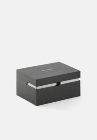 Boîte noire texturée avec une fermeture magnétique, dotée d'un intérieur blanc et d'un logo en relief minimaliste sur le dessus. Les dimensions sont compactes et rectangulaires.