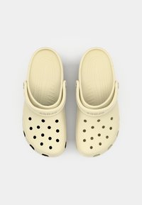Sabots beige clair avec des trous de ventilation circulaires noirs, semelle intérieure texturée et une sangle au talon ajustable, portant le nom de la marque sur le côté.
