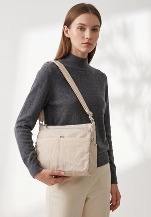 Donna che indossa un dolcevita grigio scuro e pantaloni chiari, che tiene una borsa a tracolla beige con più scomparti con cerniera.