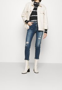 Chaqueta de mezclilla blanca, suéter de rayas negras y blancas, jeans azules desgastados y botas de tobillo blancas con tacón ancho.