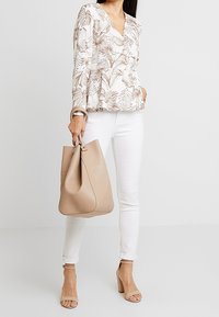 Borsa a tracolla in pelle beige con una texture morbida e liscia e un design curvo. Indossata con una blusa floreale, jeans bianchi e sandali con tacco beige.