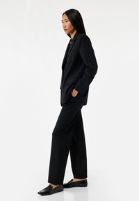 Svart skräddarsydd kostym med skarp slag, vida byxor och fickor. Parat med svarta loafers, mot en enkel vit bakgrund.