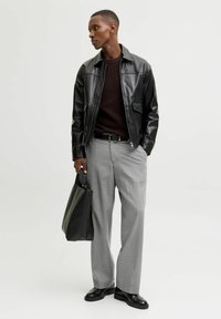 Mustaline nahktagi tumepruunist sviitrist, hallidega ruuduliste pükstega ja mustade kingadega. Käes must käekott.