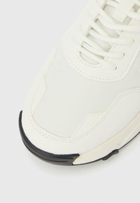 Λευκό αθλητικό παπούτσι με υφαντό υλικό στο πάνω μέρος, περίτεχνο ράψιμο και μαύρο καουτσούκ κάλυμμα στα δάχτυλα. Ορατά τα κορδόνια και η σχεδίαση των οφθαλμών.