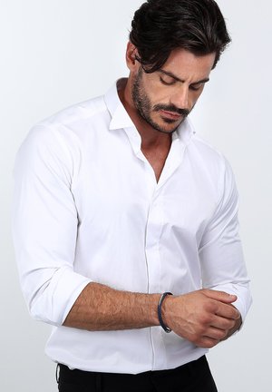 Chemise blanche à manches longues avec des poignets retroussés, texture lisse et col classique. Un bracelet sombre est visible au poignet gauche.