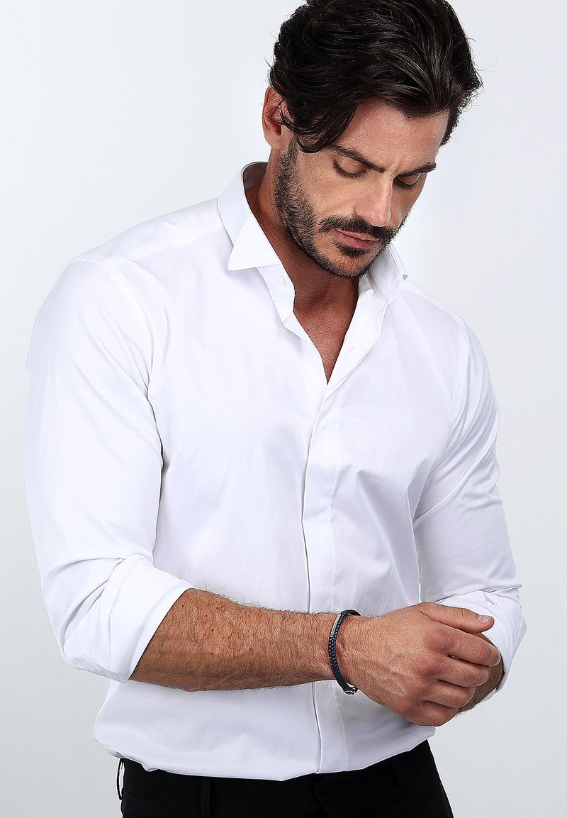 Vit långärmad skjorta med uppvikt manschetter, slät textur och en konventionell krage. Ett mörkt handledsarmband syns på vänster handled.