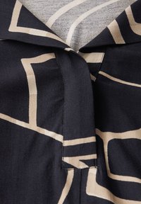 Tessuto nero con motivo geometrico crema, caratterizzato da un colletto e una patta verticale. La superficie appare liscia con una texture sottile.