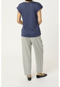 Persona in piedi di spalle che indossa una maglietta a maniche corte blu navy, pantaloni larghi grigio chiaro e scarpe basse nere su uno sfondo bianco.