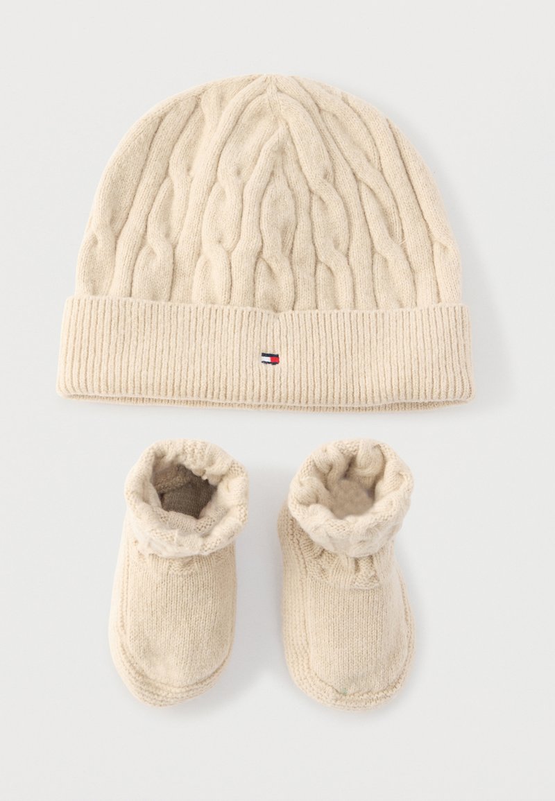 Flødefarvet strikket hat med kabelmønster og lille logo over matchende bløde strikkede babyfutter på hvid baggrund.