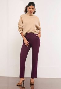 Pulover bej, din tricot moale, cu guler înalt, purtat împreună cu pantaloni ajustați, de culoare mov închis, cu talie medie. Modelul poartă pantofi cu vârf ascuțit, de culoare maronie, stând în fața unui fundal neutru.