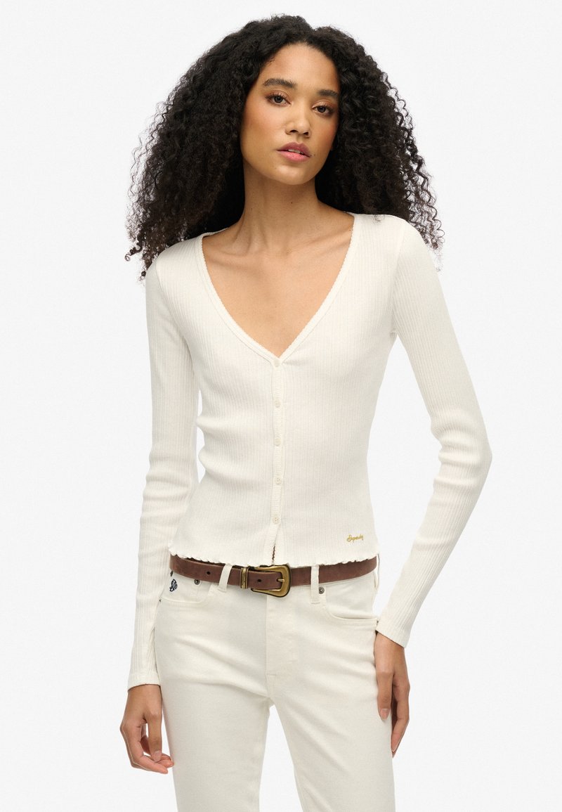 Geribde witte cardigan met een diepe V-hals, lange mouwen en knoopsluiting, met een ruwe zoom en een klein gouden logo-detail op de taille.