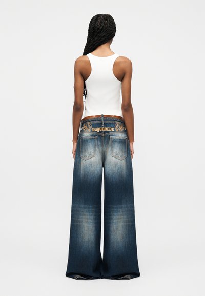 Frau mit langem, gedrehtem Haar, die ein weißes Tanktop und weite, verwaschene blaue Jeans mit besticktem "DSQUARED2"-Bund trägt, von hinten gesehen.
