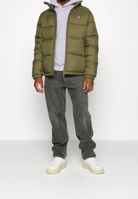 Oliwkowo zielona puchowa kurtka z zamkiem błyskawicznym, ściągacze na rękawach; noszona na jasnofioletowej bluzie z kapturem. Szare dżinsy i białe trampki dopełniają stylizację.