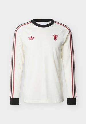 Balta krekla ar garām piedurknēm, melnā apkakle un aproces, sarkanas un melnas svītras uz piedurknēm, sarkans Adidas logotips un sarkans lauva ģerbonis uz krūtīm.