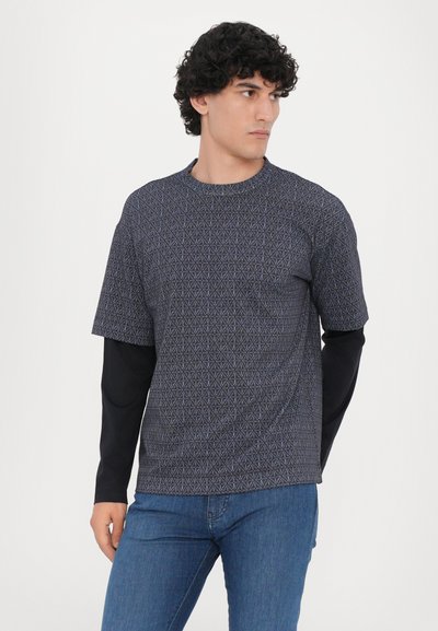 Tumšzils ar rakstu T-krekls ar īsām piedurknēm un ģeometriskām dizainparaugiem, uzvilkts pār melnu krekla ar garām piedurknēm. Zilās džinses noslēdz apģērbu.