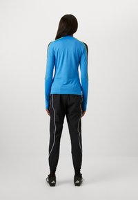 Blaues, langärmliges Sportoberteil mit schwarzen Schulterakzenten und gelben Details, kombiniert mit schwarzen Jogginghosen mit weißen Seitenstreifen.
