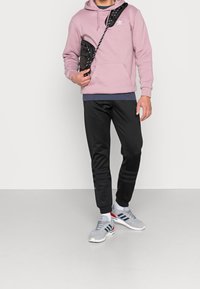 Ljusrosa huvtröja, svarta sweatpants med blå detaljer, grå löparskor med röda och blå ränder, samt en svart axelremsväska.