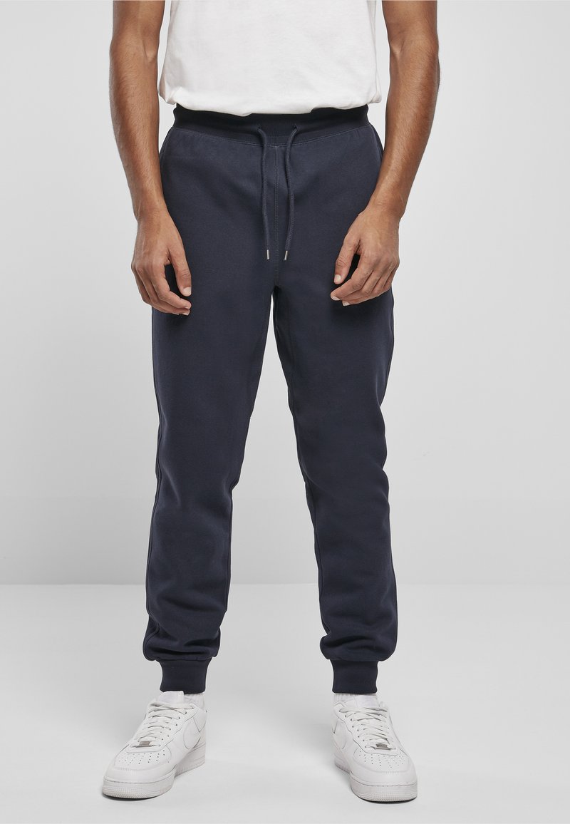 Mann trägt marineblaue Jogginghosen mit Kordelzug am Bund, kombiniert mit weißen Turnschuhen und einem weißen T-Shirt, steht auf grauem Boden.