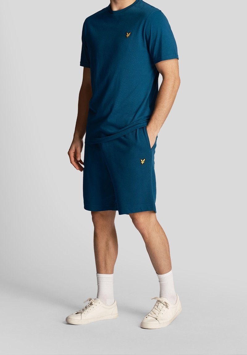 T-shirt teal et shorts assortis en tissu doux ; présente un petit logo doré sur la poitrine et sur les shorts. Des baskets blanches et des chaussettes de cheville complètent la tenue.
