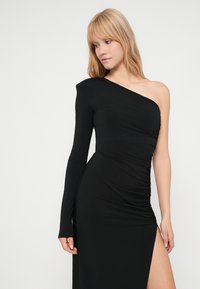 Crna haljina s jednim ramenom, izrađena od glatke tkanine, s uskim siluetom, naborima na boku i prorezom do natkoljenice.