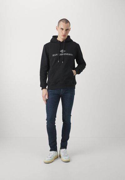 Črna majica s kapuco z logotipom "HAN COHENHAVN", sprednji žep, kapuca z vrvico; v kombinaciji z temno modrimi jeans hlačami in belimi supergami z rumenimi podplati.