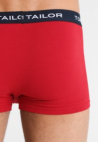 Rote Baumwollboxershorts mit schwarzem elastischem Bund, der mit weißem Text "TOM TAILOR" in einem wiederkehrenden Muster versehen ist. Glatte Textur und enganliegendes Design.