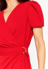 Rotes Wickelkleid aus glattem Stoff, mit einem tiefen V-Ausschnitt und kurzen Puffärmeln. Seitliche Raffung betont durch ein goldenes Ringdetail.