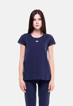 Giovane donna con capelli lisci e scuri che indossa una maglietta blu navy a maniche corte e pantaloni abbinati su sfondo bianco.