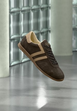 Zapatilla deportiva GANT marrón y beige con cordones flotando sobre un suelo gris reflectante, frente a una pared de bloques de vidrio y pilares de hormigón.