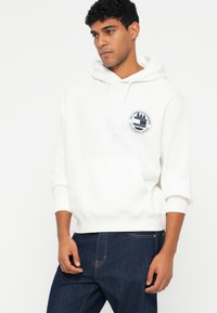 Baltas džemperis su kišene priekyje, apdailintas apskritu tamsiai mėlynu logotipu krūtinėje. Minkšta tekstūra, raukšlėtos rankovių ir apačios kraštai, derinamas su tamsiai mėlynomis džinsais.