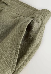 Gros plan sur un pantalon en tissu texturé vert olive, mettant en avant la ceinture élastique et l'ouverture de la poche latérale.
