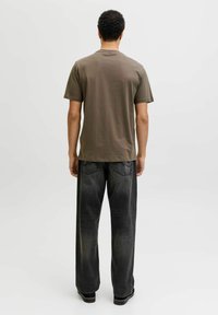 Hombre de pie de espaldas, vistiendo una camiseta de manga corta marrón, pantalones vaqueros negros descoloridos y zapatos negros sobre un fondo blanco liso.