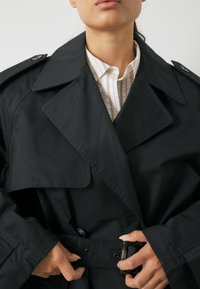 Czarny płaszcz trench z szerokimi klapami, podwójnymi guzikami i paskiem w talii. Teksturowany materiał ma delikatny połysk. Nosi się go na jasnej, paskowanej koszuli.