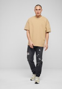 Camiseta de algodón beige de corte oversized con mangas cortas, combinada con vaqueros negros desgastados con rotos y detalles, y zapatillas deportivas de color claro.