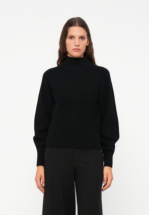 Zwarte geribbelde coltrui met een ontspannen pasvorm, lange mouwen en een gestructureerd oppervlak. Gecombineerd met zwarte wijde pantalons.