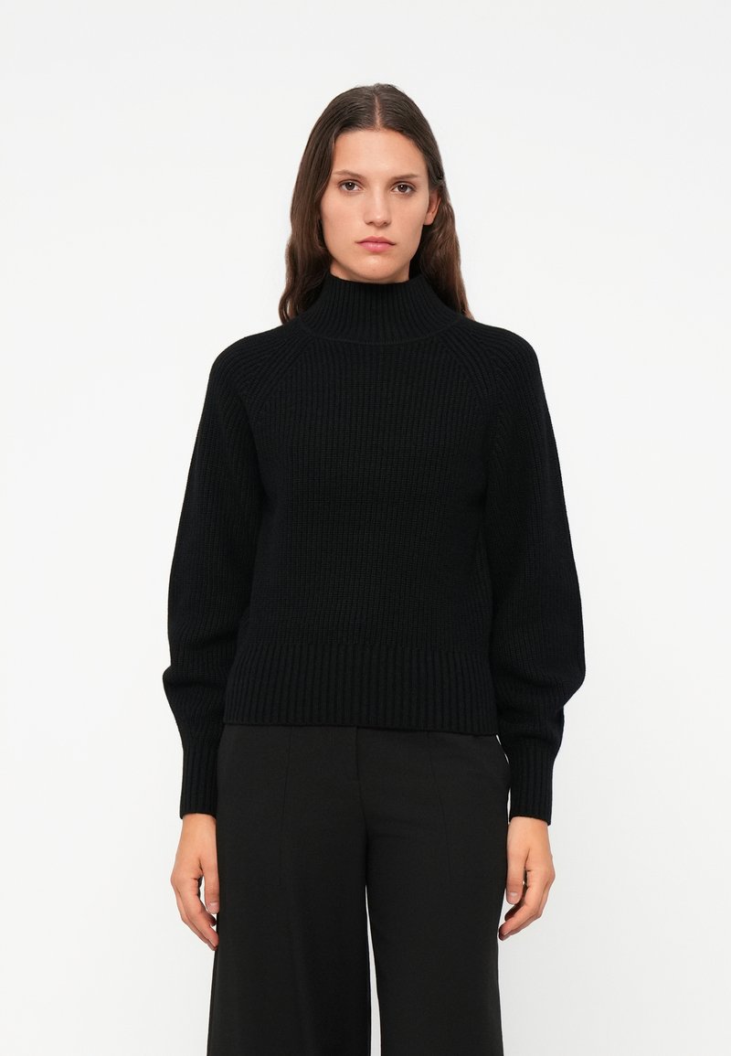 Pull à col roulé noir côtelé avec une coupe décontractée, des manches longues et une surface texturée. Associé à un pantalon large noir.