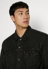 Jacă din denim negru cu guler clasic, buzunare frontale și nasturi metalici. Are o textură decolorată și un detaliu mic cu logo roșu.